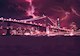 The photograph depicts a large suspension bridge, illuminated at night against an urban skyline, during a lightning storm. 